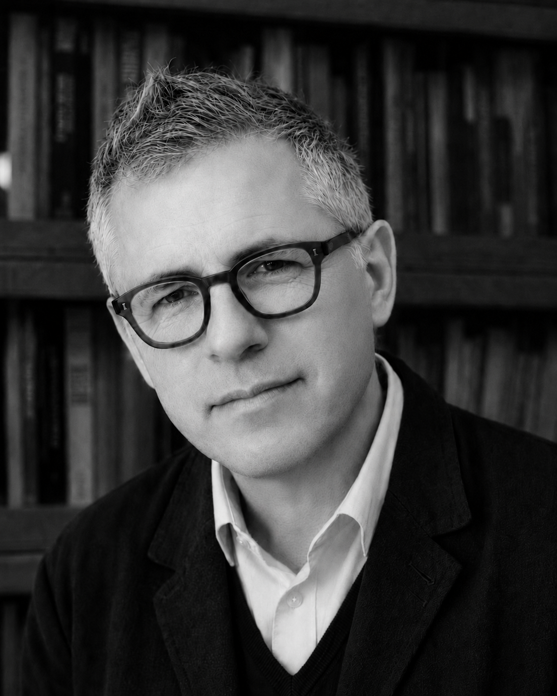 Man in square glasses lookin gat the camera, with bookshelf out of focus behind him. In black and white