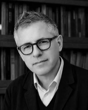 Man in square glasses lookin gat the camera, with bookshelf out of focus behind him. In black and white