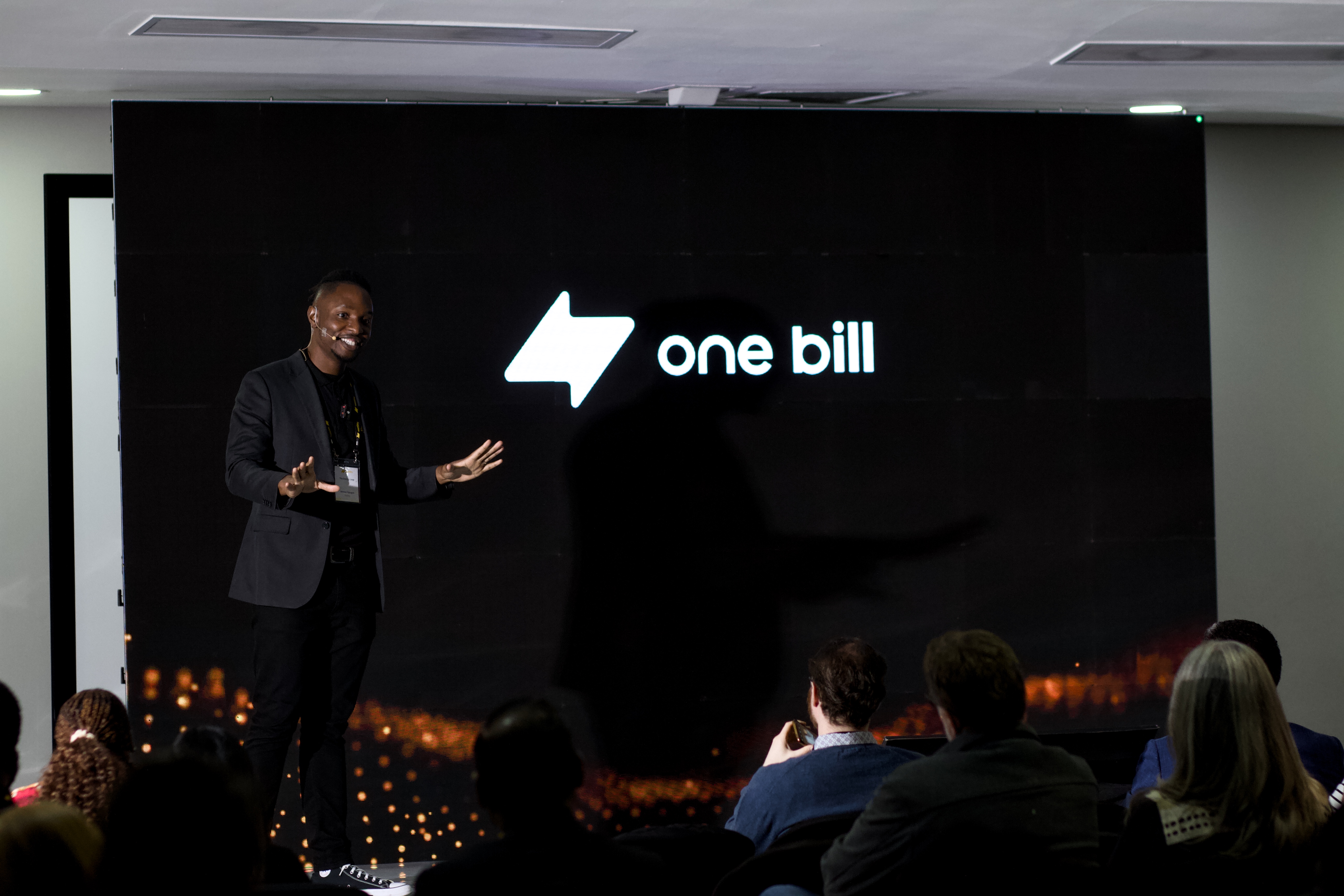 Man in blazer stands on a stage smiling towards the audience, in front of black screen which reads "one bill" in white writing.
