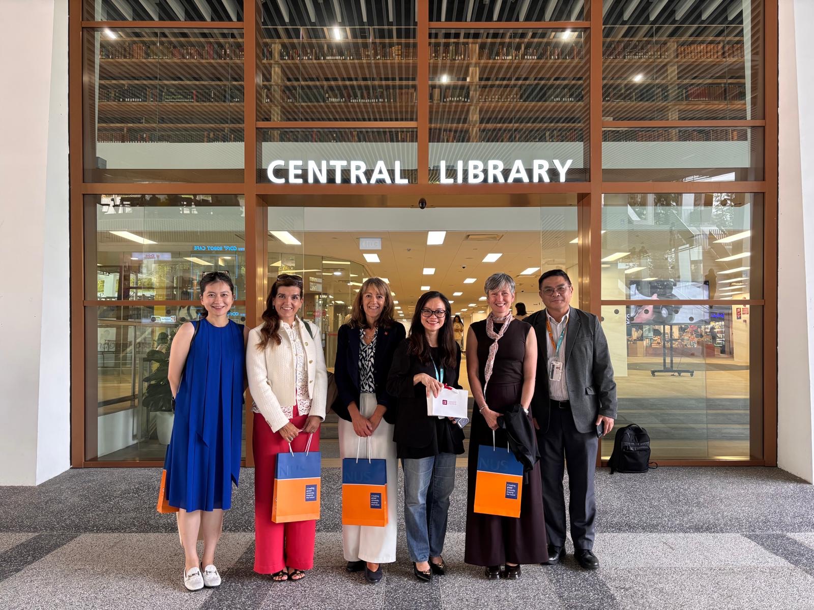 Six individuals posing in front of NUS's Central Library holding NUS themed gift bags.