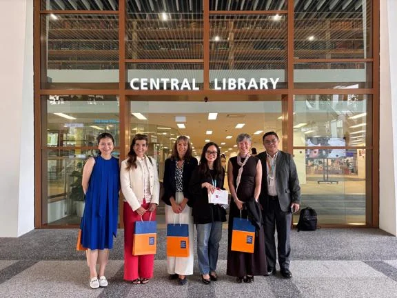 Six individuals posing in front of NUS's Central Library holding NUS themed gift bags.