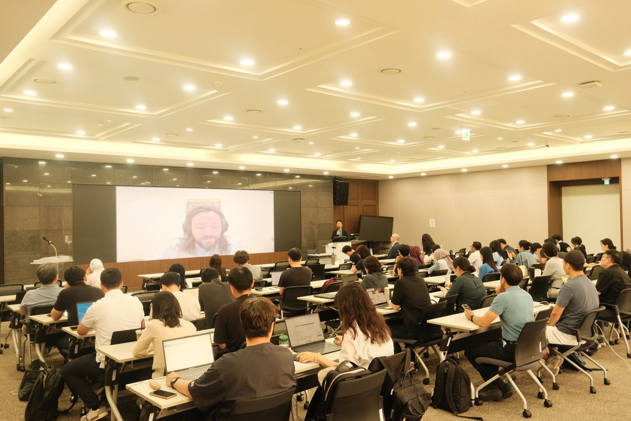 A photo of the workshop in a classroom at the KDI School, with attendees sitting a desks watching a speaker in person and on the screen at the front of the room. 