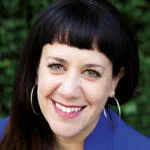 Yael is smiling at the camera, wearing a blue jacket and hoop earrings.