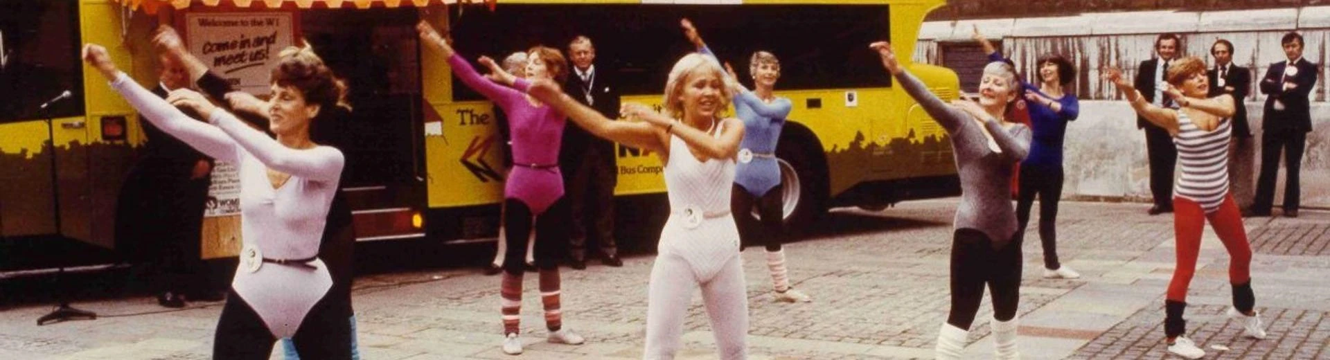 A group of people doing aerobics in the street. There is a bus behind them with Women in the Community written on it.