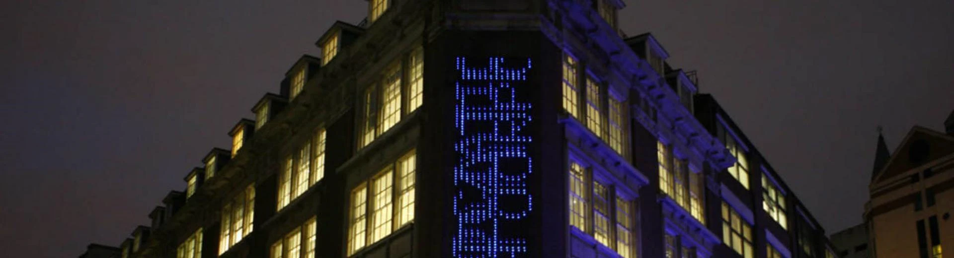 An outside view of LSE Library at night which includes the artwork Bluerain - an installation where searches from the catalogue scroll down from top to bottom on top of each other using blue LED lights.