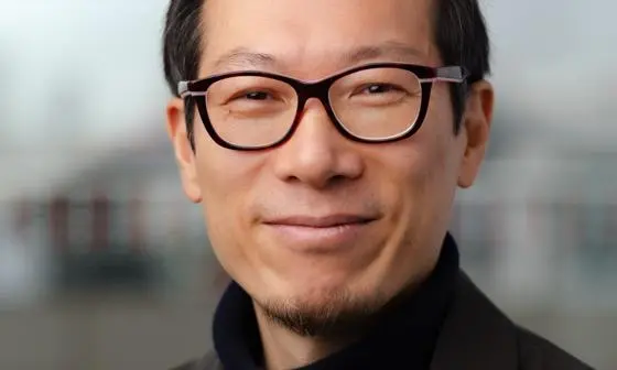 Hyun Bang Shin looking to camera wearing black glasses and black suit.