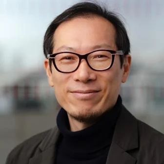 Hyun Bang Shin looking to camera wearing black glasses and black suit.
