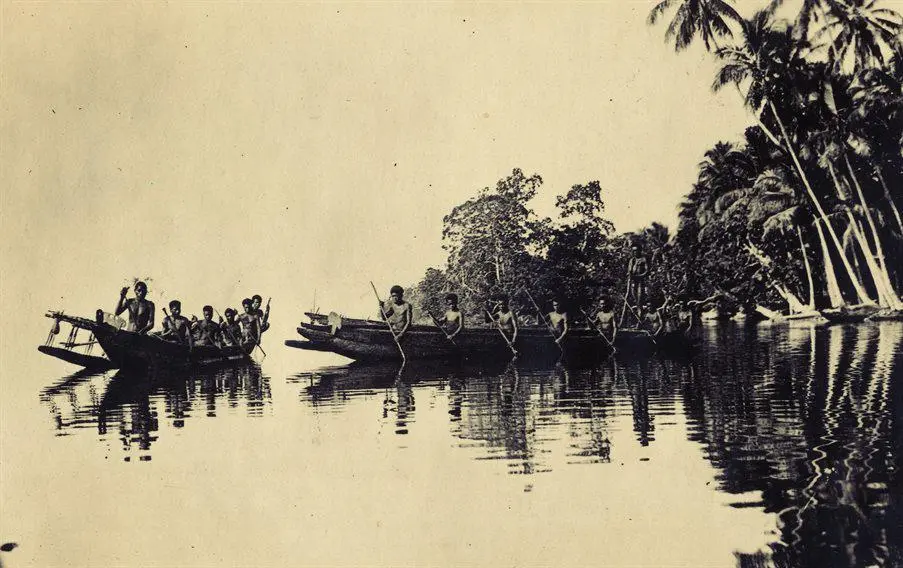 A photograph across water of two canoes being sailed by Trobriand islanders. There are trees in the background.