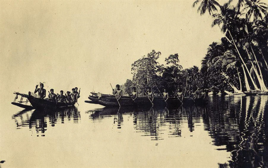 A photograph across water of two canoes being sailed by Trobriand islanders. There are trees in the background.