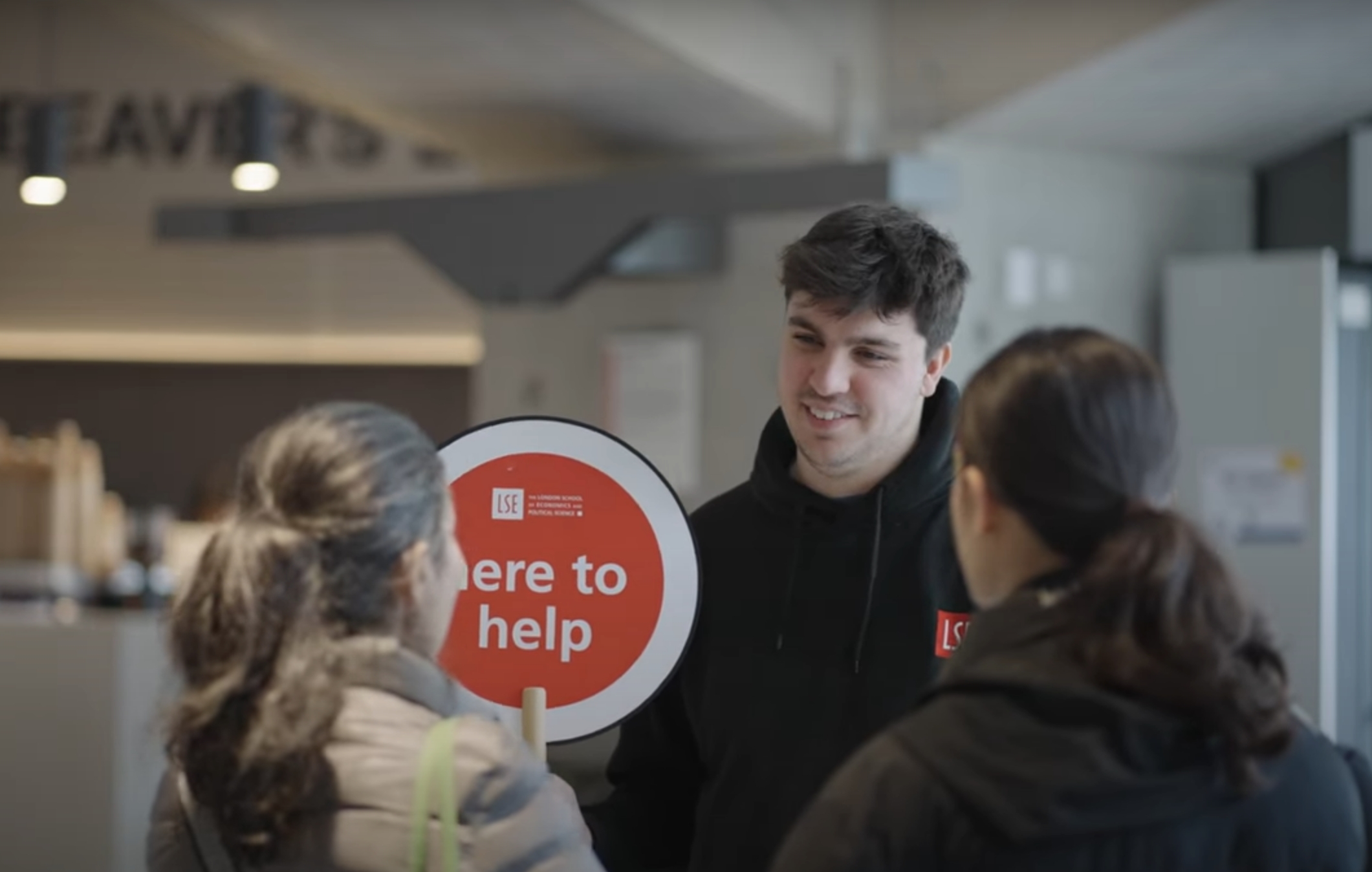 A Student Ambassador holding a 'here to help' sign talking to visitors.