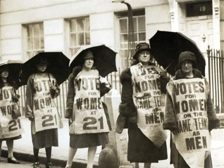 A group of suffrage women marching with umbrellas and placards