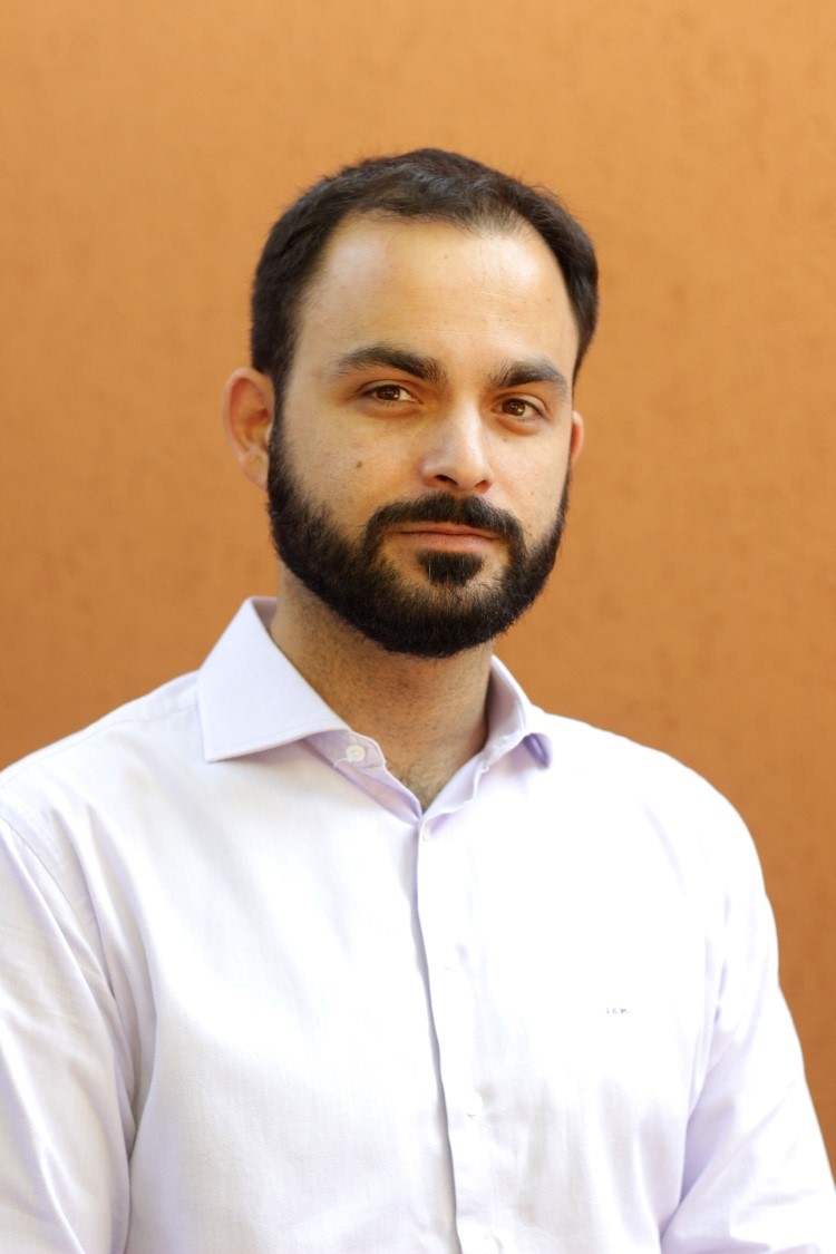 Ivan Marqus is facing the camera. He is wearing a light-colored button-up shirt standing against a yellow background.