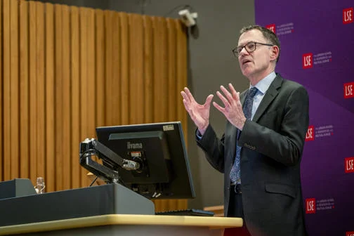 Man in a suits stood in front of an LSE banner and behind a lectern delivering a lecture