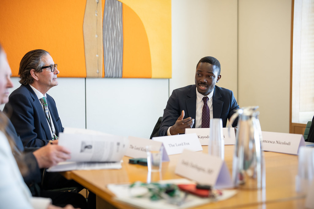 Several people are sat around a table with name labels in front of them, listening to a man in a suit gesturing with his hands.