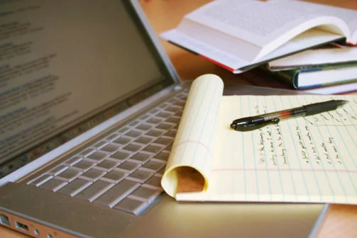 Laptop computer with open books in the background, and a pen resting on a yellow legal pad with writing on it.