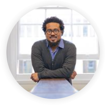 Upper body front profile image of Lasana.  He is leaning on a tabletop and has crossed arms.  He's wearing a grey jumper over a mid blue collared shirt.  Lasana has black curly hair, short to medium length and is wearing round spectacles.