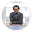 Upper body front profile image of Lasana. He is leaning on a tabletop and has crossed arms. He's wearing a grey jumper over a mid blue collared shirt. Lasana has black curly hair, short to medium length and is wearing round spectacles.