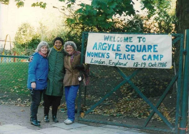 Three women stood at a gate reading 'Welcome to Argyle Square Women's Peace Camp, London, England 19-19 Oct 1986.