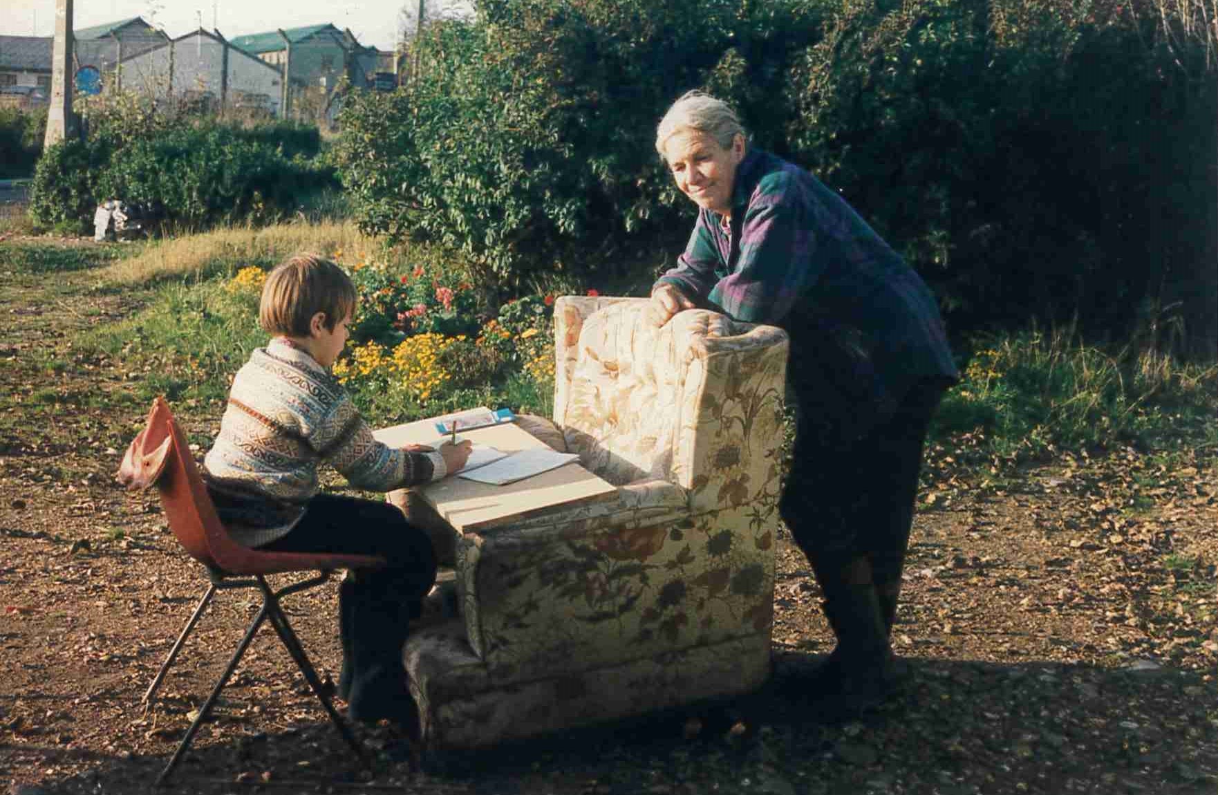 A child sat at a makeshift desk using a board and an armchair with an adult stood leaning on the armchair 