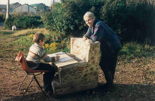 A child sat at a makeshift desk using a board and an armchair with an adult stood leaning on the armchair