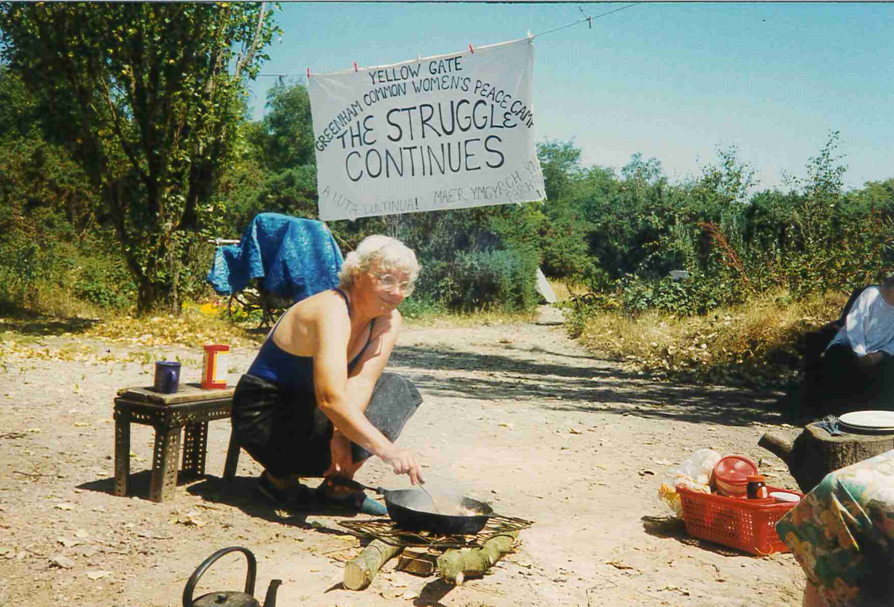 A persn sat outside cooking on a fire with a banner hanging behind.