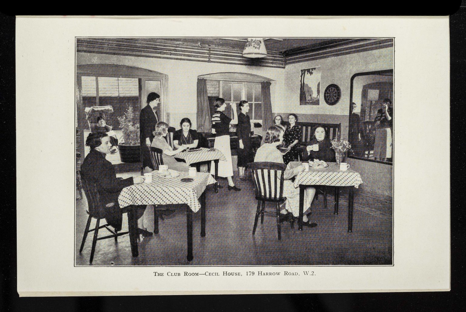 A club room with people sat at tables and others playing darts.