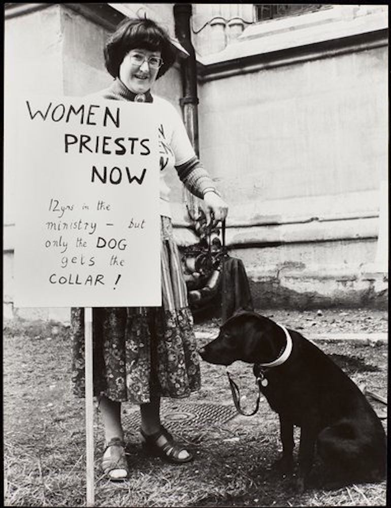 A woman stood with a placard reading "Women Priests Now" and a dog.