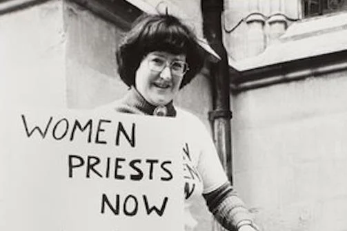 A woman stood with a placard reading "Women Priests Now" and a dog.
