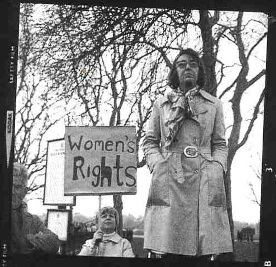 A woman in a trenchcoat in front of another holding a Women's Rights placard. 