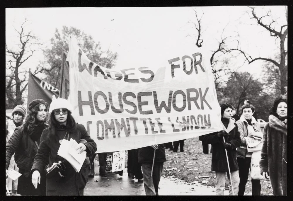A group of women marching holding a banner reading 'WAGES FOR HOUSEWORK COMMITTEE LONDON'