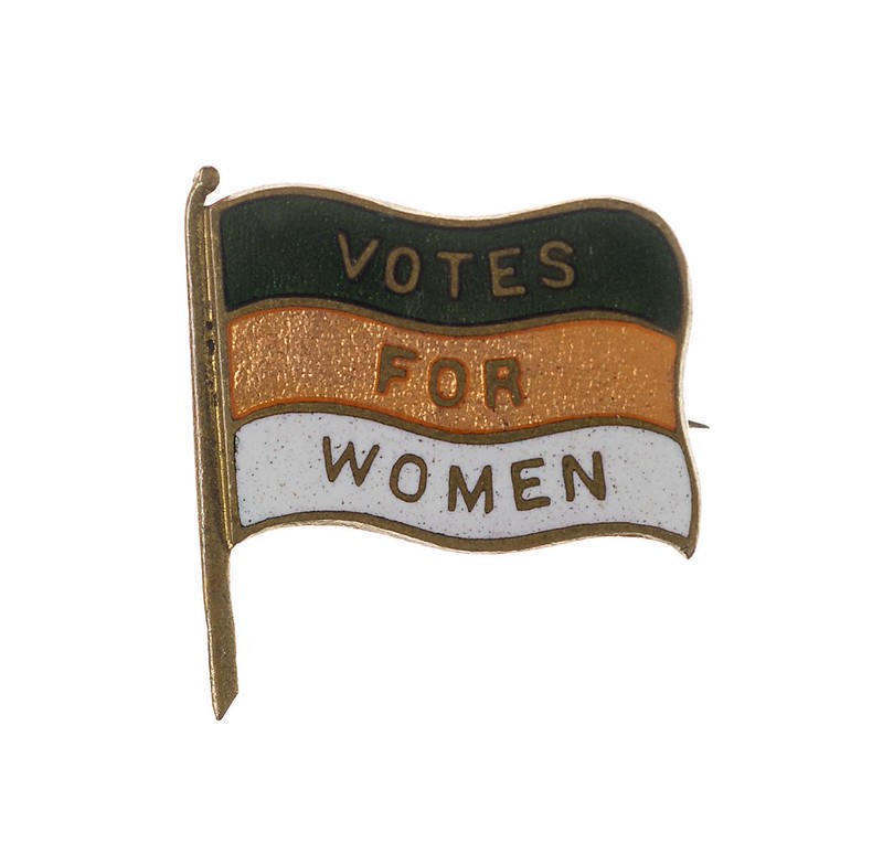 A green, gold and white Women's Freedom League badge with Votes For Women written on it. 