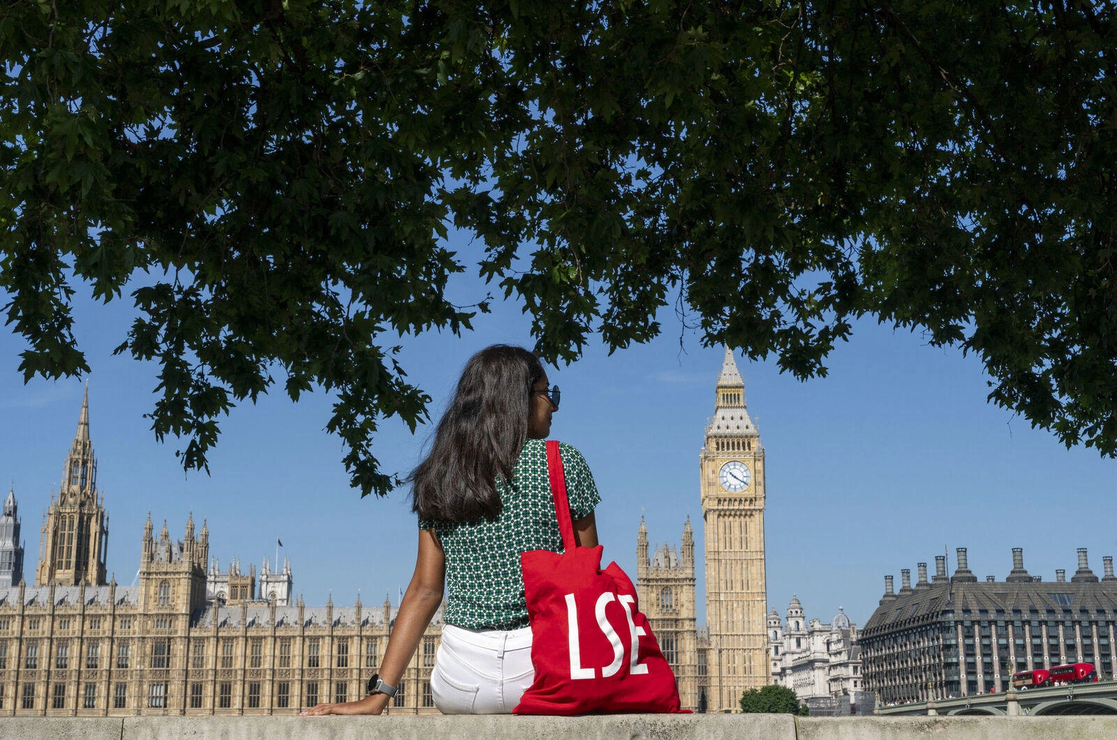 An LSE student with a red LSE tote bag sits by the river Thames overlooking Westminster on a sunny day