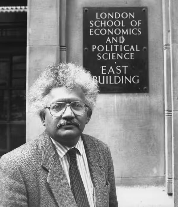 A black and white photograph of Lord Desai in front of a sign for LSE, East Building. 