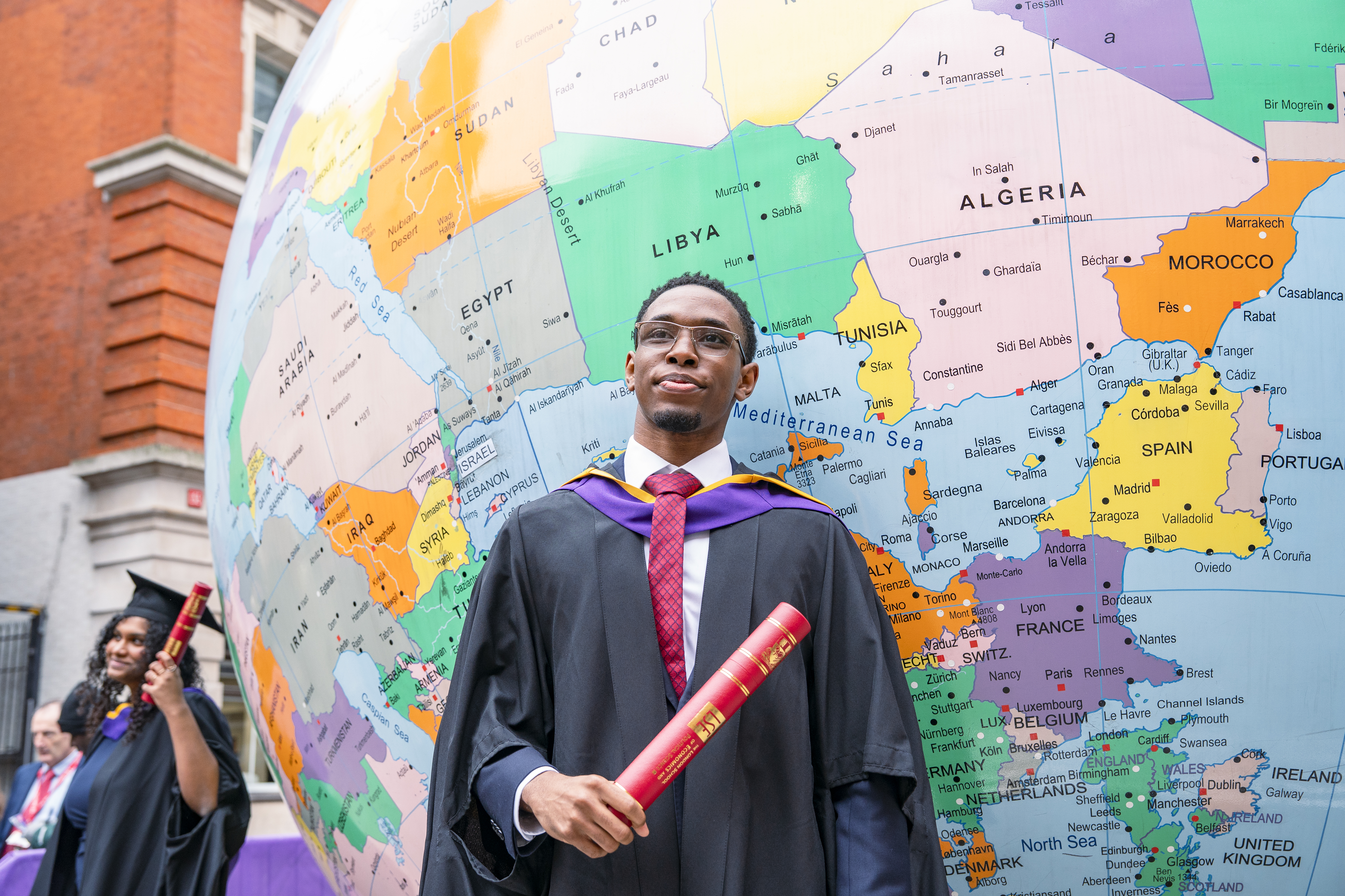 Graduate holding a diploma stood in front of upside down globe statue on campus.
