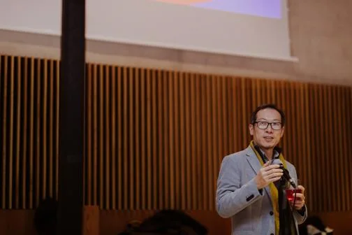 Hyun Bang Shin speaking into a microphone at an event in the Great Hall, Marshall Building