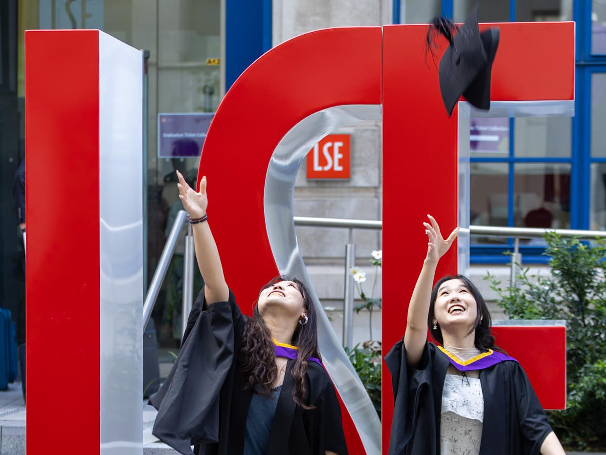 Two female graduates throwing their mortar boards in the air in front of large red L S E letters