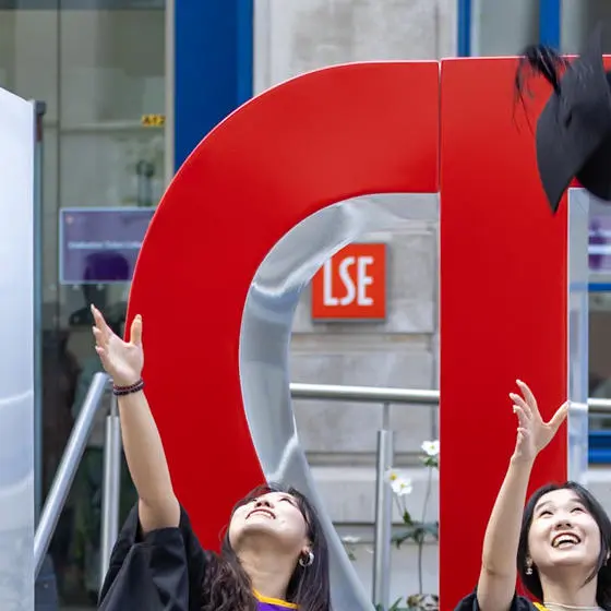 Two female graduates throwing their mortar boards in the air in front of large red L S E letters