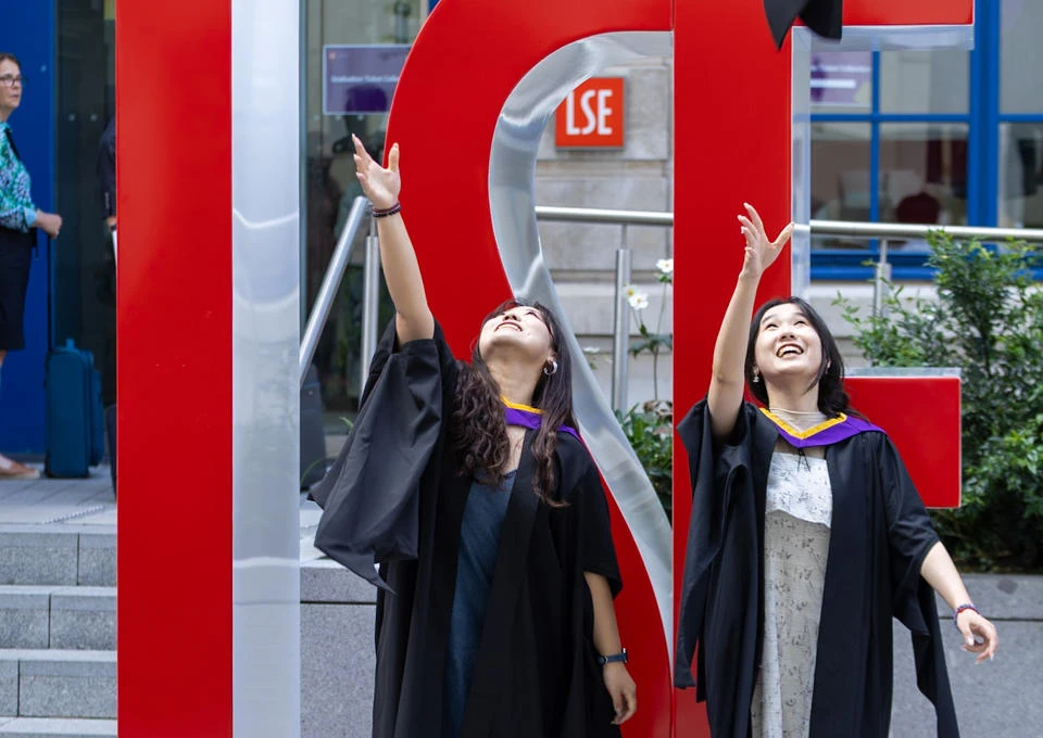 Two female graduates throwing their mortar boards in the air in front of large red L S E letters