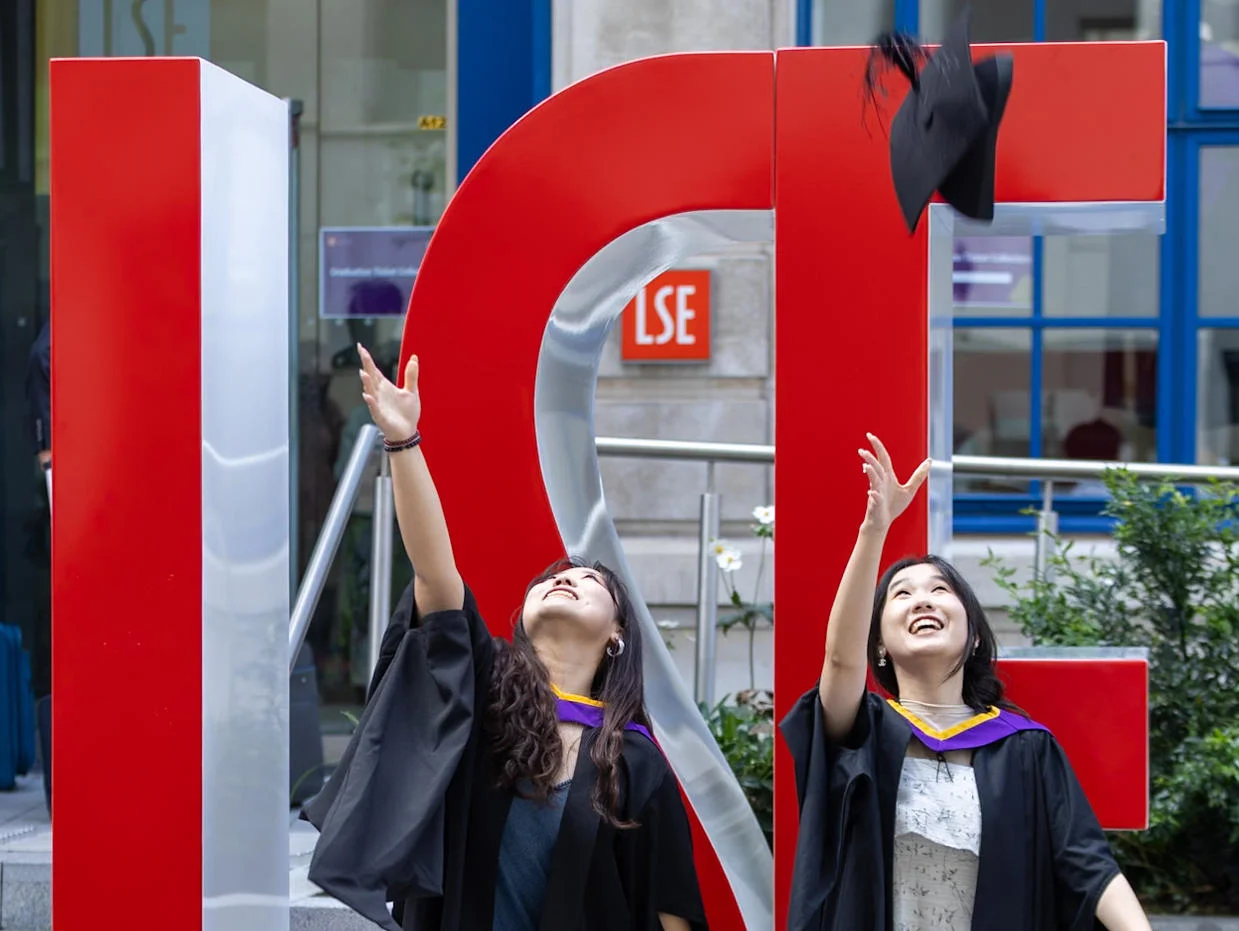 Two female graduates throwing their mortar boards in the air in front of large red L S E letters