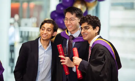 Three students wearing graduation robes, smiling and posing with their degree scrolls.