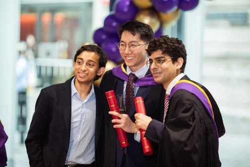 Three students wearing graduation robes, smiling and posing with their degree scrolls.