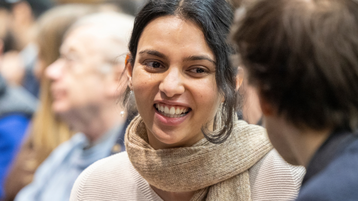 Woman smiling and talking with another person in a crowded indoor setting.