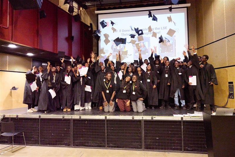 A large group wearing graduation robes, standing on a stage, throwing their mortar boards in the air.