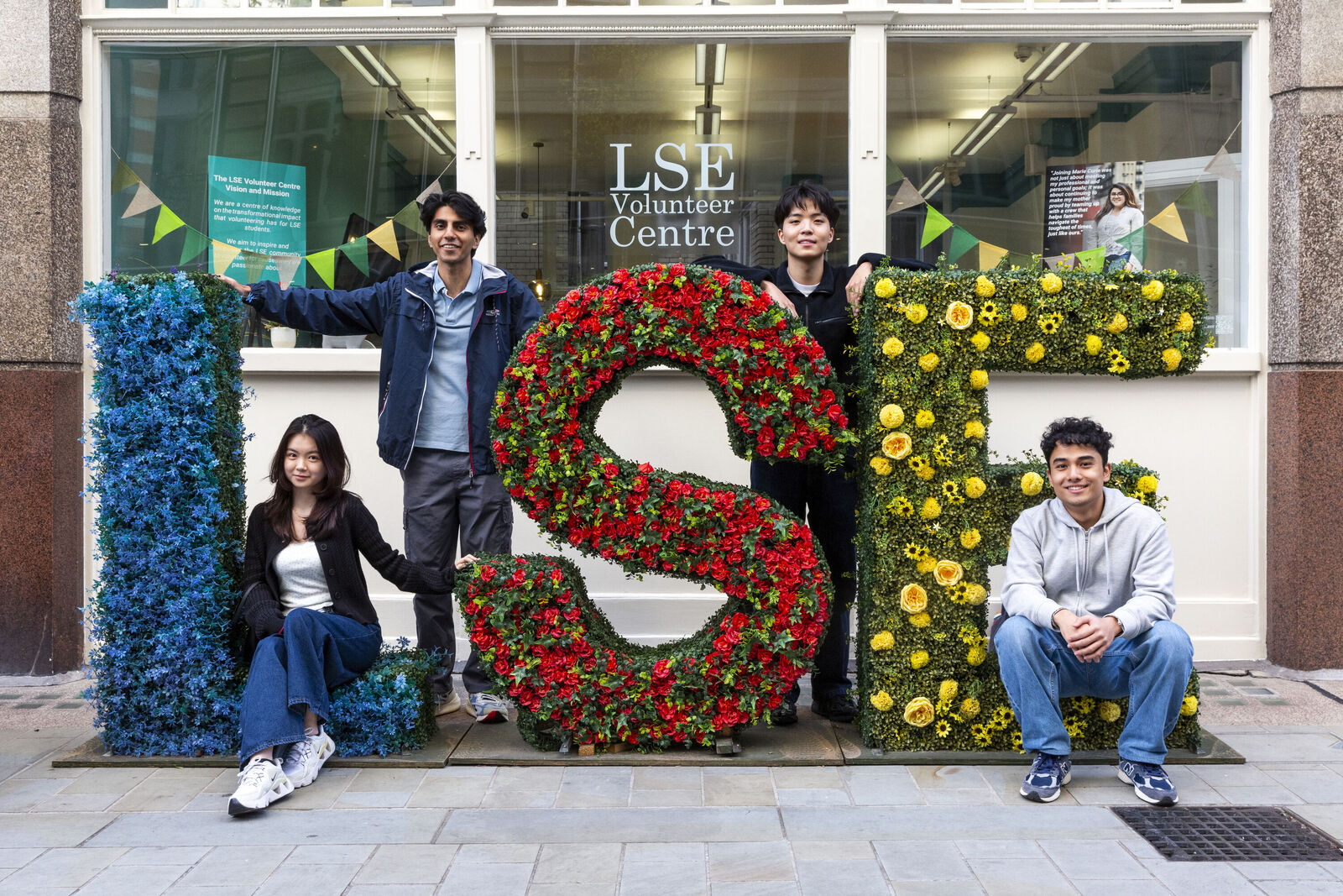 Students on LSE campus sitting and standing next to flower display which spells 'LSE'