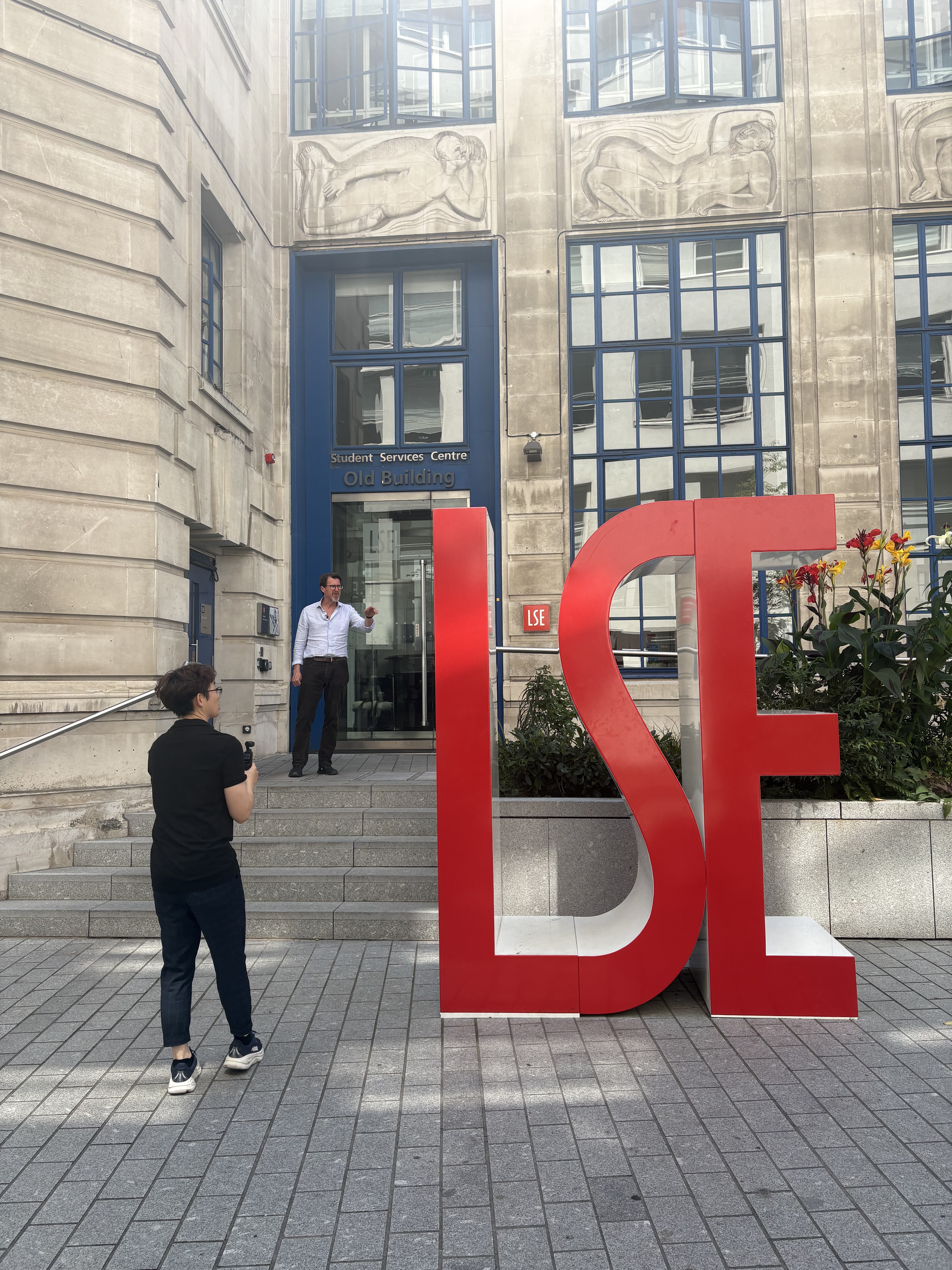 huge LSE letters, with two men stood next to them. One holds a camera, filming the other