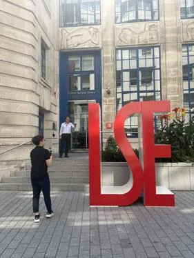 huge LSE letters, with two men stood next to them. One holds a camera, filming the other