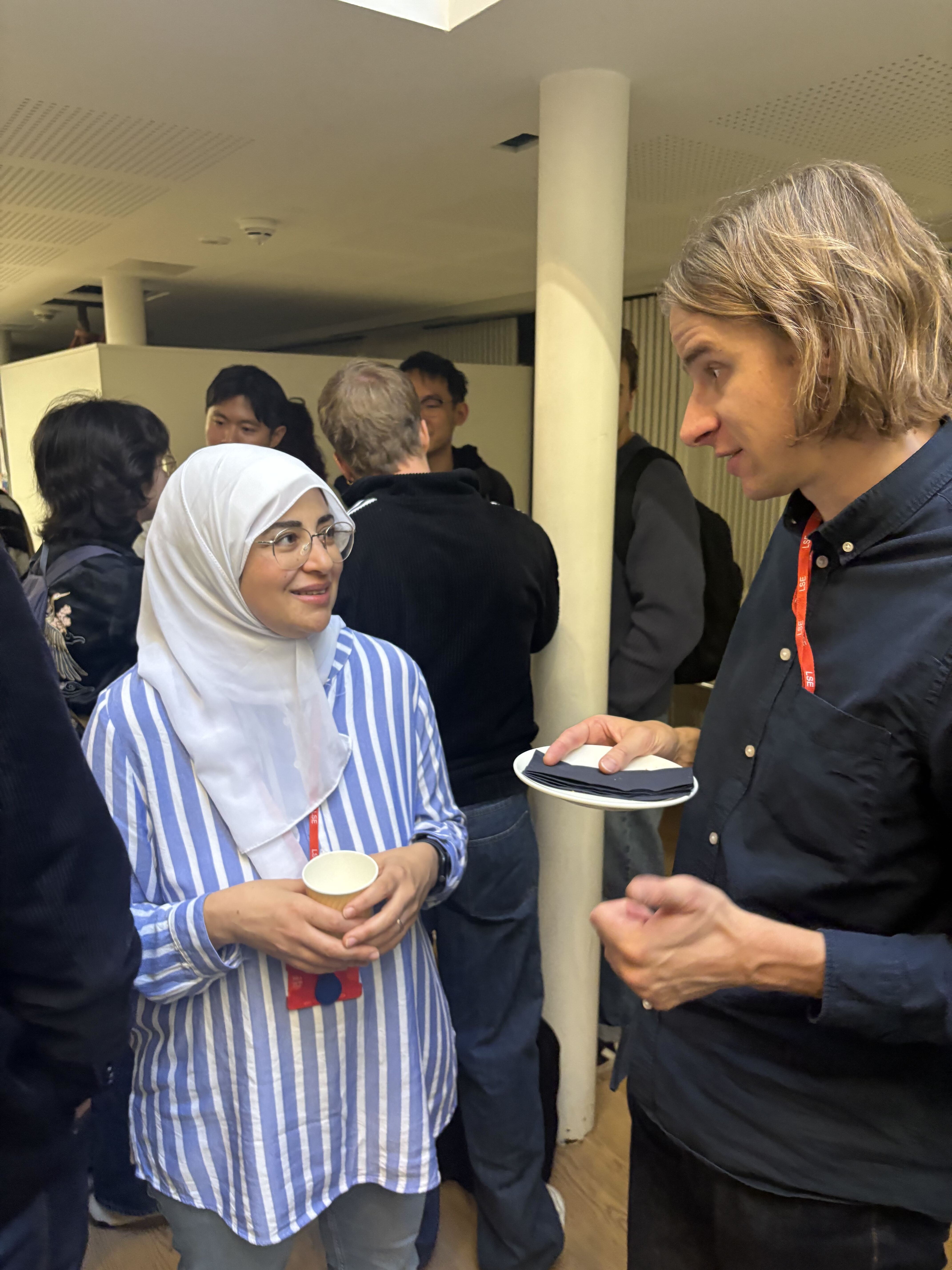 A man and woman stand and converse at an indoor gathering, each holding a plate of food, with other people in the background.