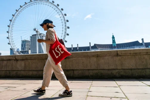 Woman walking opposite the London eye carrying a red LSE bag