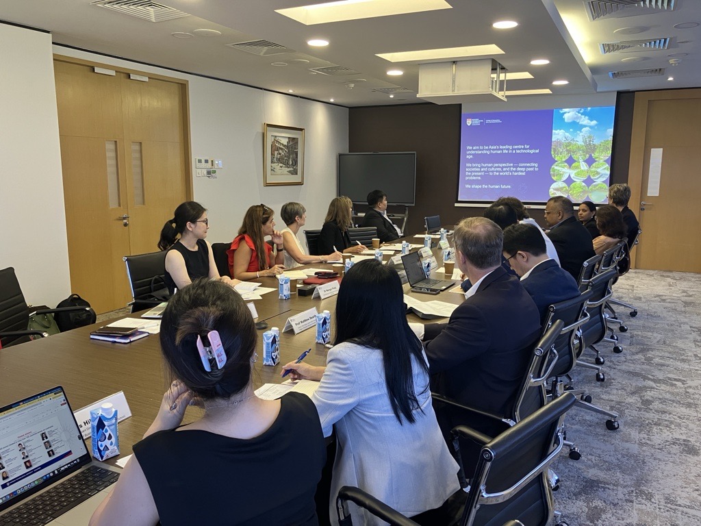 Individuals sitting at a long table in a meeting room watching a presentation. 