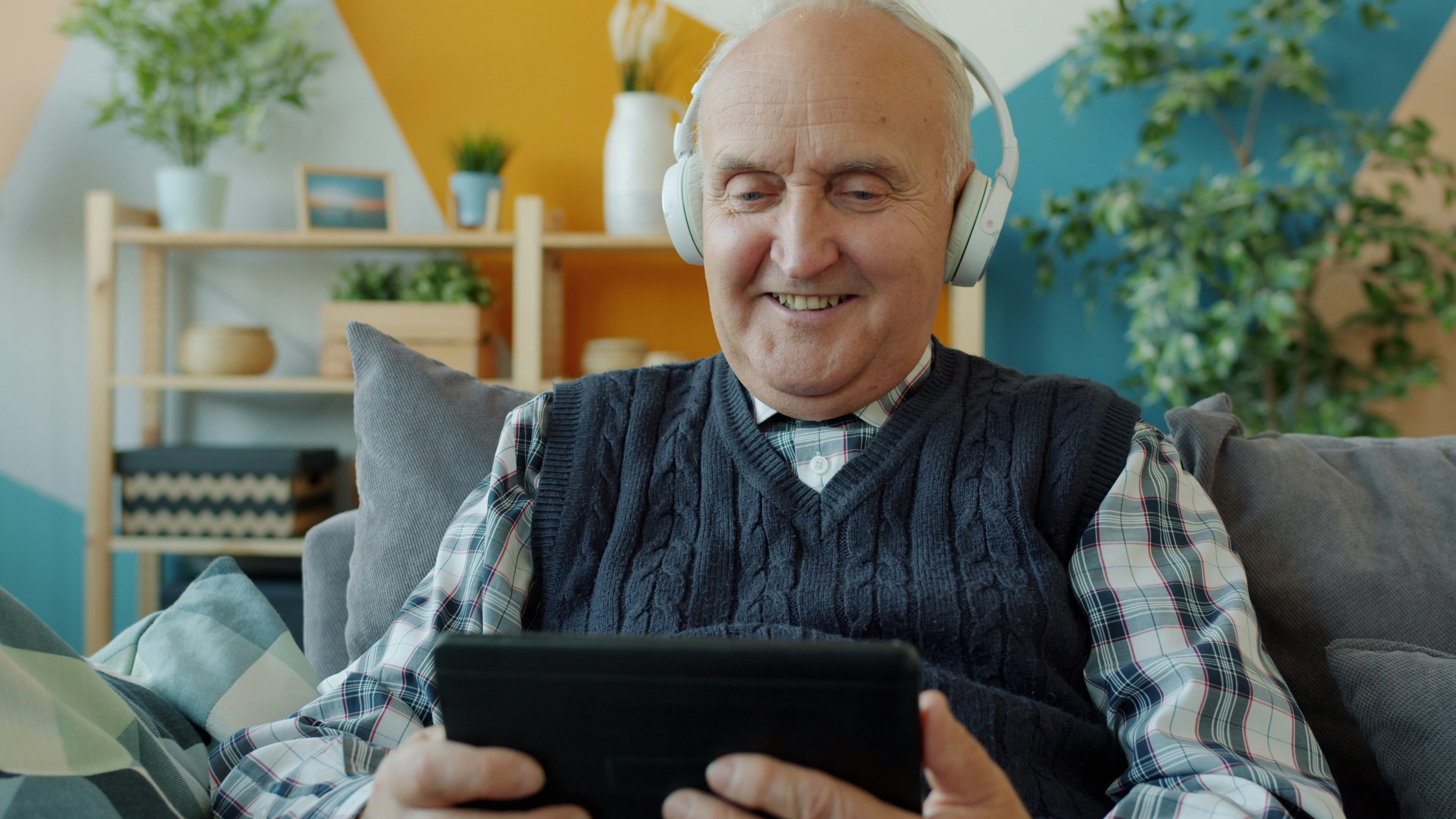 An elderly man sat on a sofa, he is reading something on a tablet whilst wearing big over ear headphones.